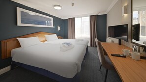 Ein Hotelzimmer mit einem großen Bett, einem Schreibtisch mit Kaffeemaschine, einem Fernseher und einem Vorhangfenster.