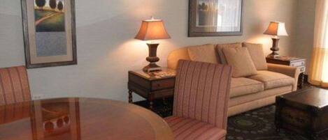 A hotel room with a round wooden dining table, a sofa, two lamps, and framed pictures on the walls.