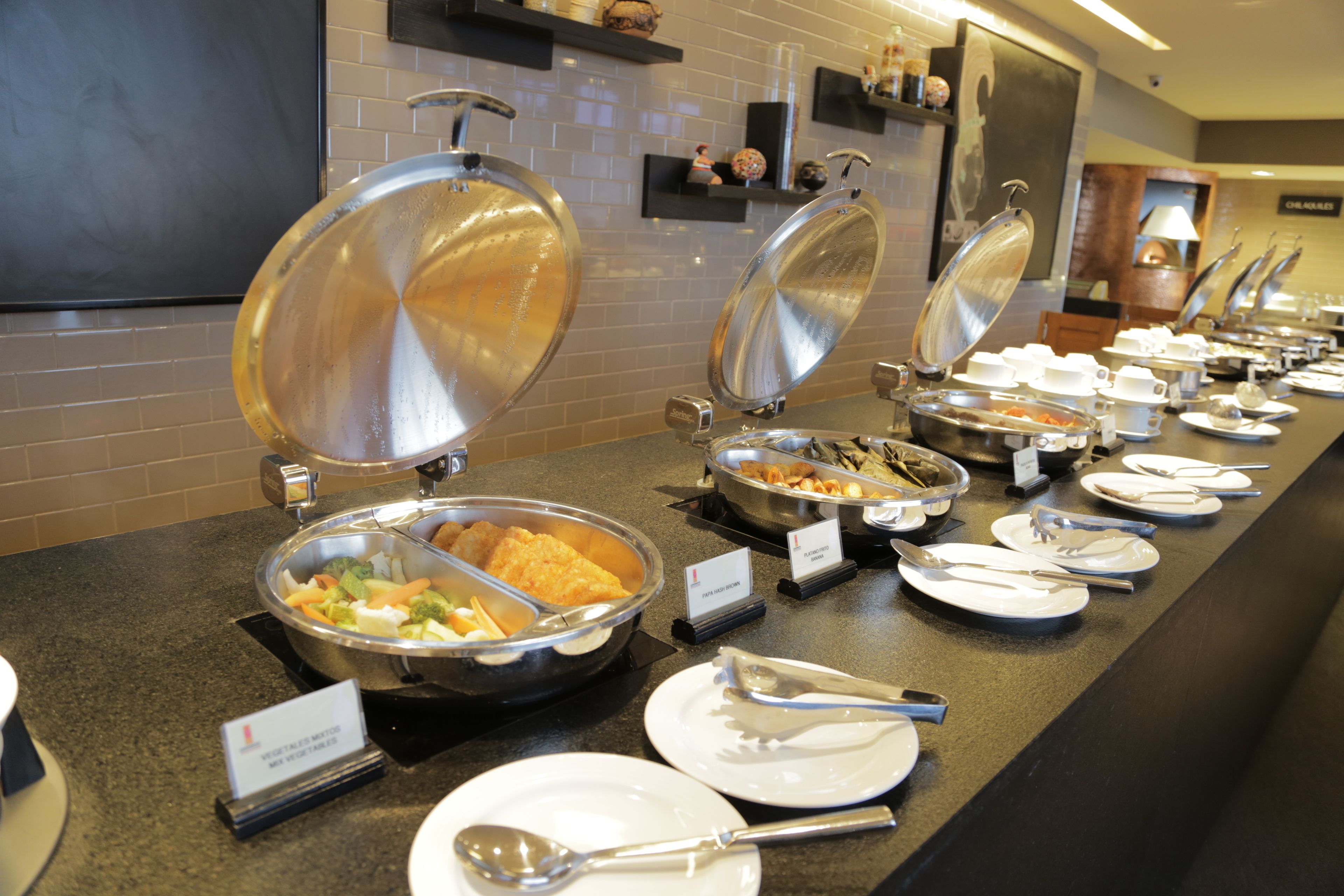Tarjolla on runsas buffet, jossa on useita ruokapisteitä, kuten salaattibaari ja lämpimien ruokien alue. Ruoanlaitto- ja tarjoiluastiat sekä lautaset on asetettu esille tiskille.