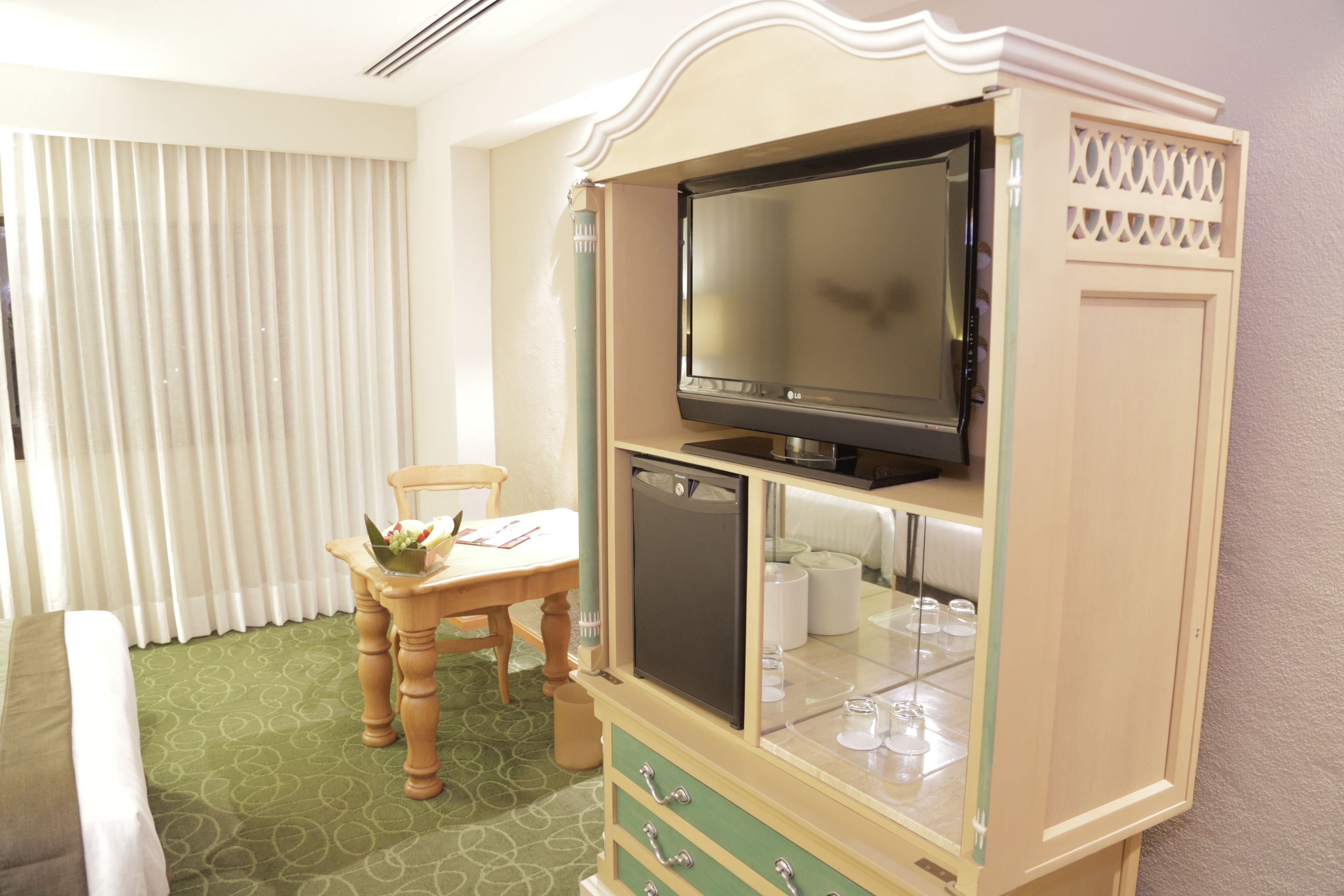 A hotel room with a television on a cabinet, a dining table with a chair, and a window with blinds.