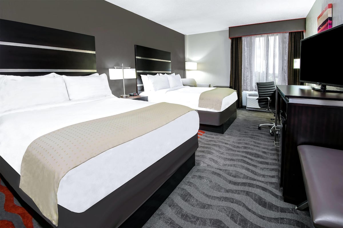 A hotel room with two beds, a desk, a chair, a TV, and a large window with curtains.