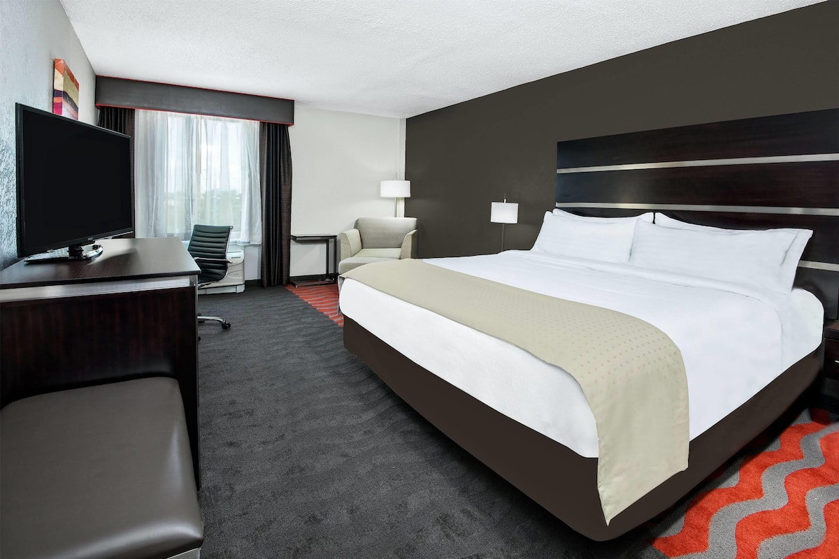 A hotel room with a large bed, a desk with a flat-screen TV, a chair, a small table, and a lamp.
