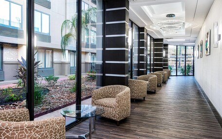 A modern hotel lobby with a long corridor, patterned chairs, a glass table, and a view of the outdoor garden.