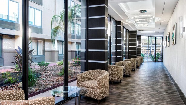A modern hotel lobby with a long corridor, patterned chairs, a glass table, and a view of the outdoor garden. - Wyndham Houston Sugarland (Houston)