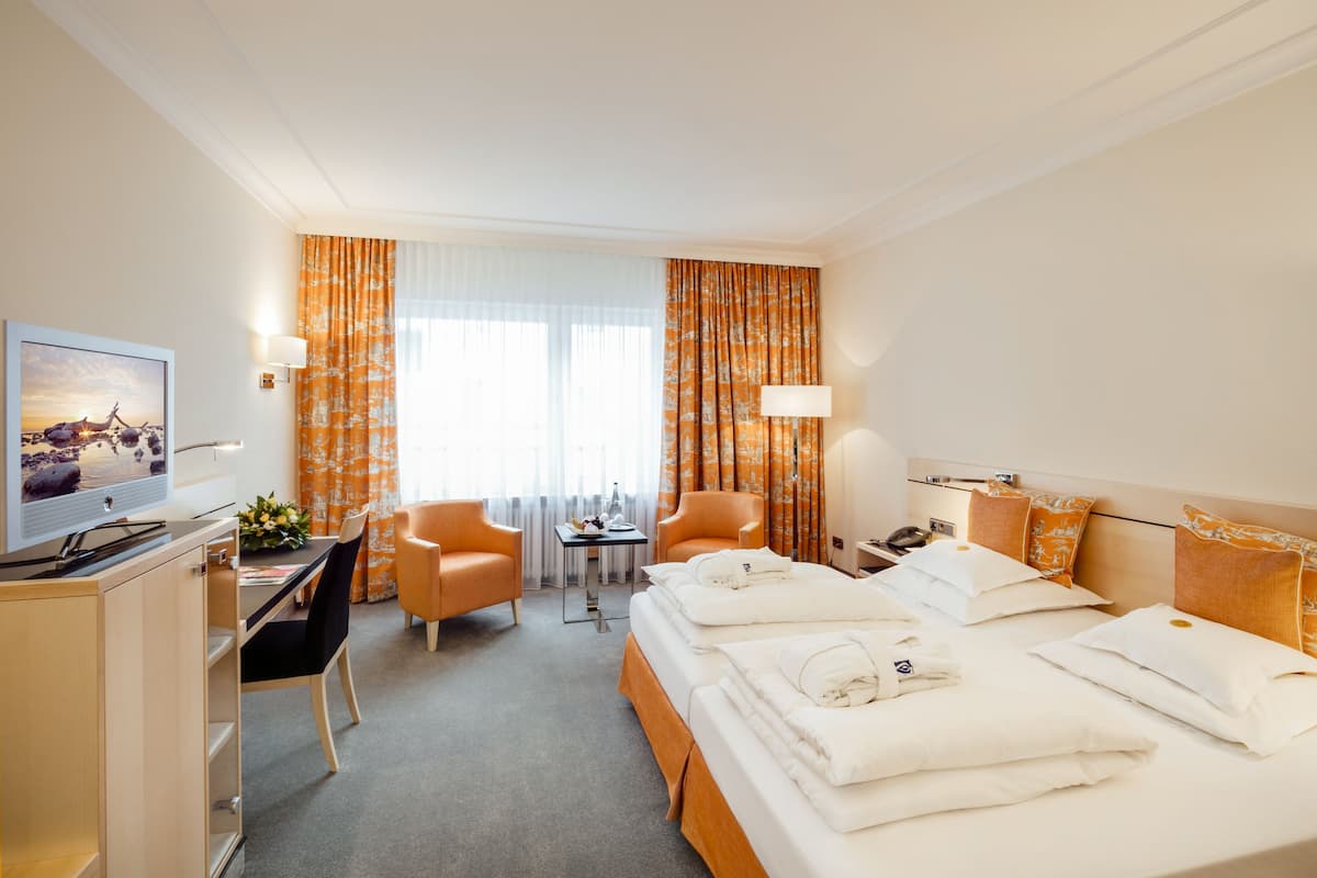 A hotel room with a bed, two chairs, a desk, a television, and a window with curtains.