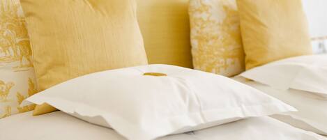 Una cama con almohadas blancas y amarillas, un cabecero sencillo y una colcha blanca con un diseño discreto.