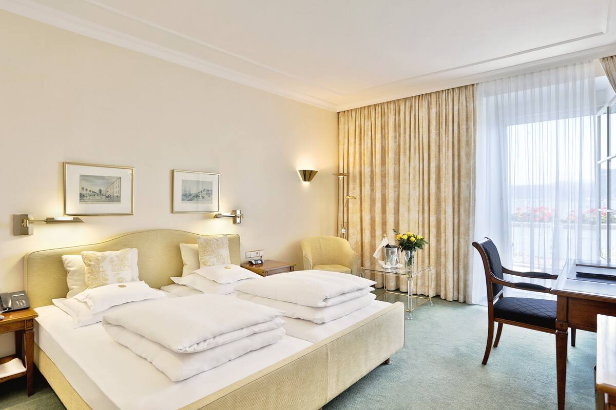 A hotel room with a large bed, two bedside tables, a desk, and a chair. There are two framed pictures on the wall, a lamp, and a window with curtains.