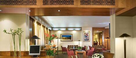 A hotel lobby with a wooden reception desk, striped chairs, a dining area with tables and chairs, and a large plant.