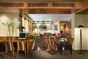 Un hall de hotel con un mostrador de recepción de madera, sillas a rayas, un comedor con mesas y sillas, y una planta grande.