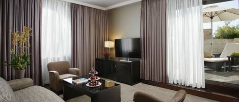 Habitación de hotel con sofá, sillones, televisión, una mesa de centro y un balcón con mobiliario exterior.
