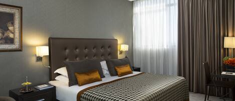 A hotel room with a large bed, two bedside tables with lamps, a desk with a chair, and a large window with curtains.