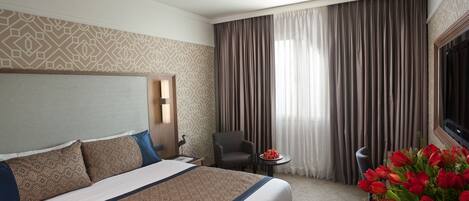 Una habitación de hotel con una cama grande, un escritorio, una silla, una mesa con fruta y un jarrón de flores.