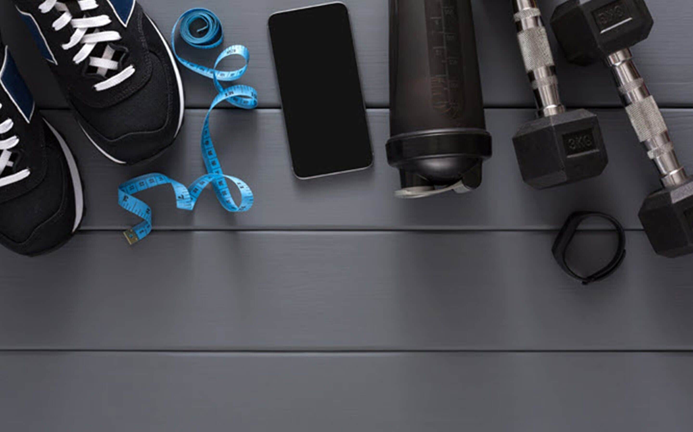 A pair of black sneakers, a blue measuring tape, a smartphone, a water bottle, and a set of dumbbells on a wooden surface.