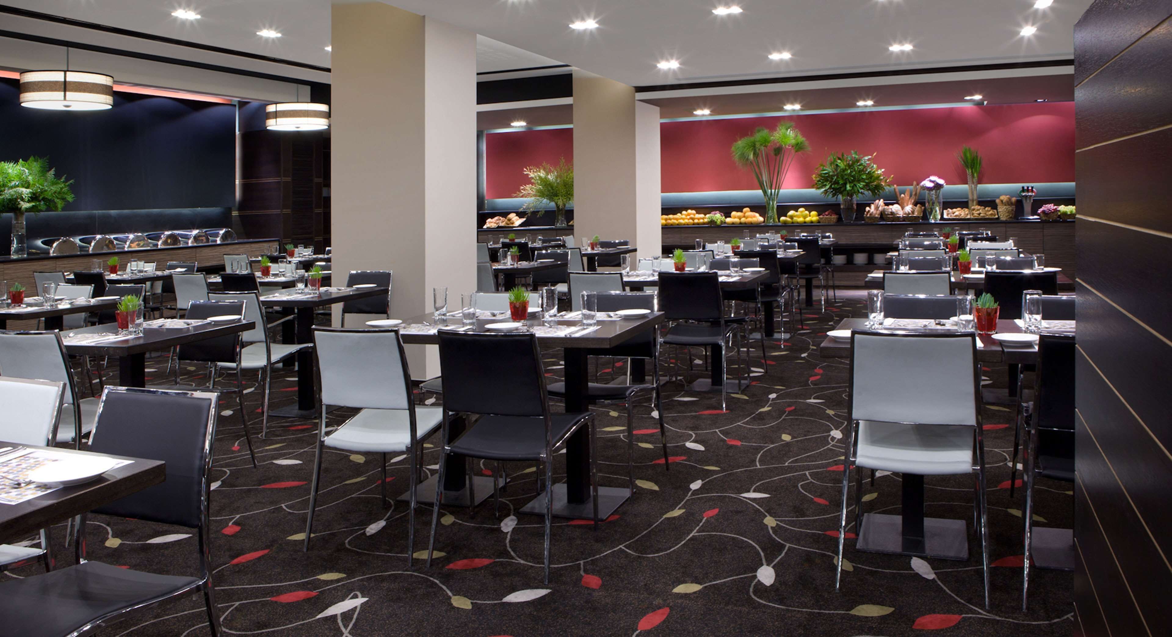 Un restaurante espacioso con varias mesas y sillas, un suelo con estampado y una pared de acento roja decorada con plantas.