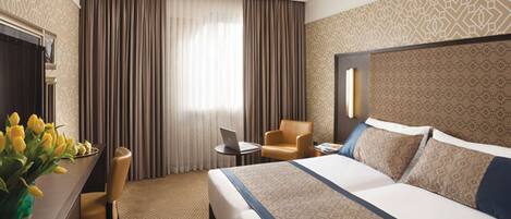 A hotel room with a large bed, a desk with a laptop, a chair, and a vase of yellow flowers.