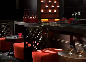 Zona de bar tenuemente iluminada con asientos tapizados en rojo, una pared decorativa con paneles iluminados y dos vasos con líquido ámbar sobre una mesa redonda de madera.