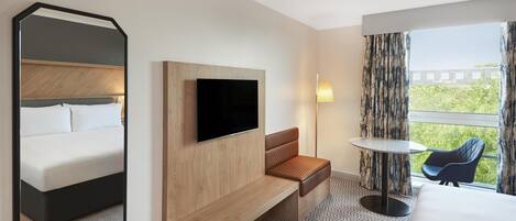 Habitación de hotel con cama, televisor, banco, mesa, silla y vistas a zonas verdes.