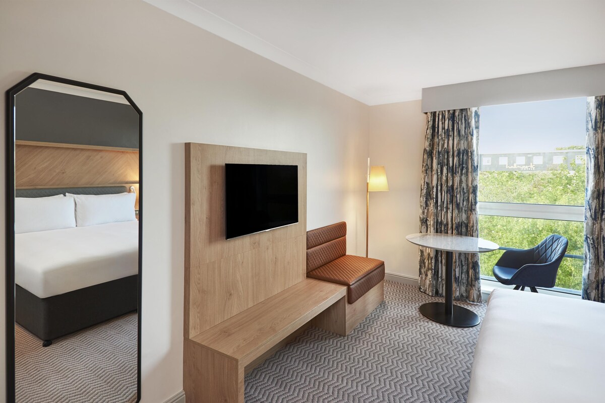 A hotel room with a bed, a TV, a bench, a table, a chair, and a view of greenery outside.