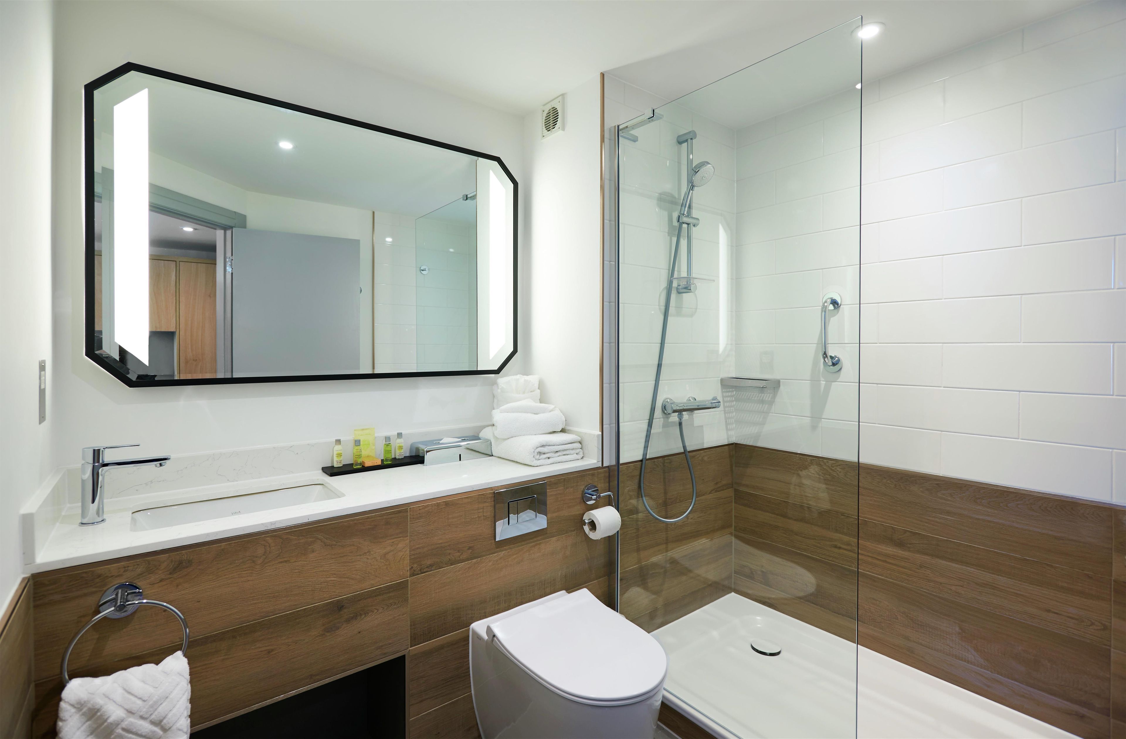 Un baño moderno con ducha envuelta en vidrio, un espejo grande y un tocador con artículos de aseo.