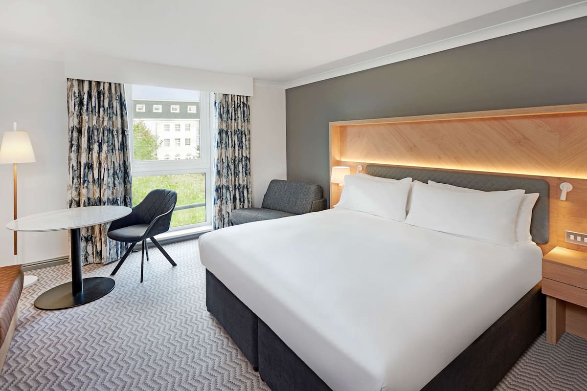 Habitación de hotel con una cama grande, un escritorio, una silla y vistas a un edificio a través del ventanal.