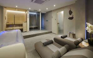 A modern spa room with a massage table, a relaxation area with sofas, a glass-enclosed shower, and a wall-mounted clock. - Hilton London Croydon (Croydon)