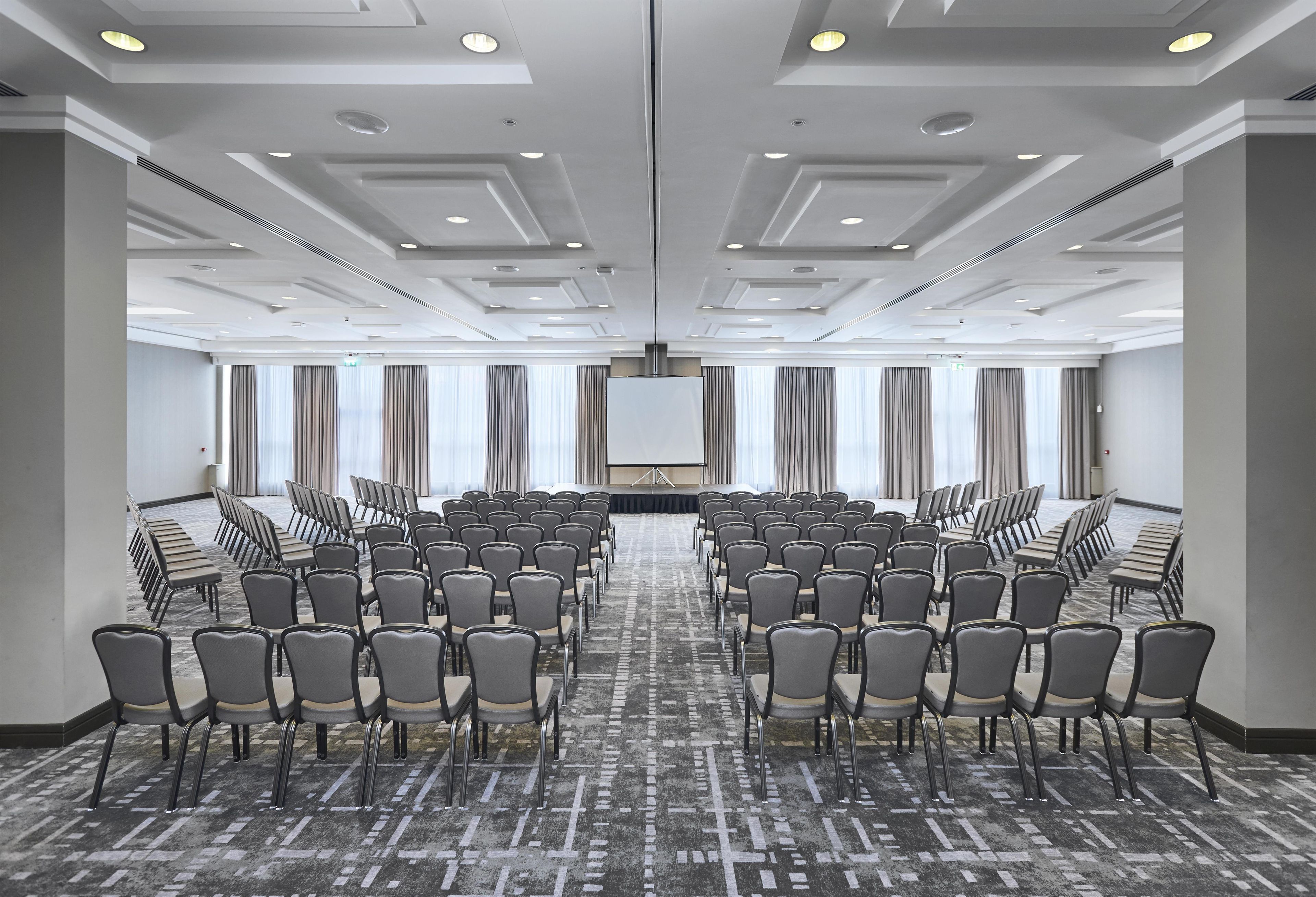 Sala de conferências com fileiras de cadeiras, uma tela grande e piso coberto de carpete.