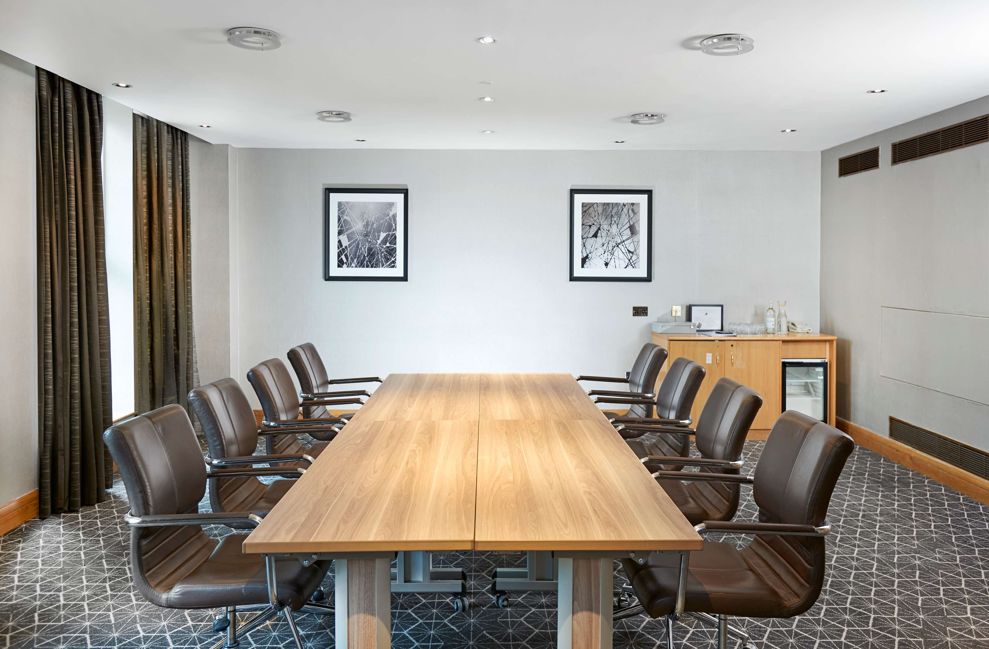 A conference room with a long wooden table, eight leather chairs, two framed abstract paintings, a small cabinet with a refrigerator, and patterned carpet.