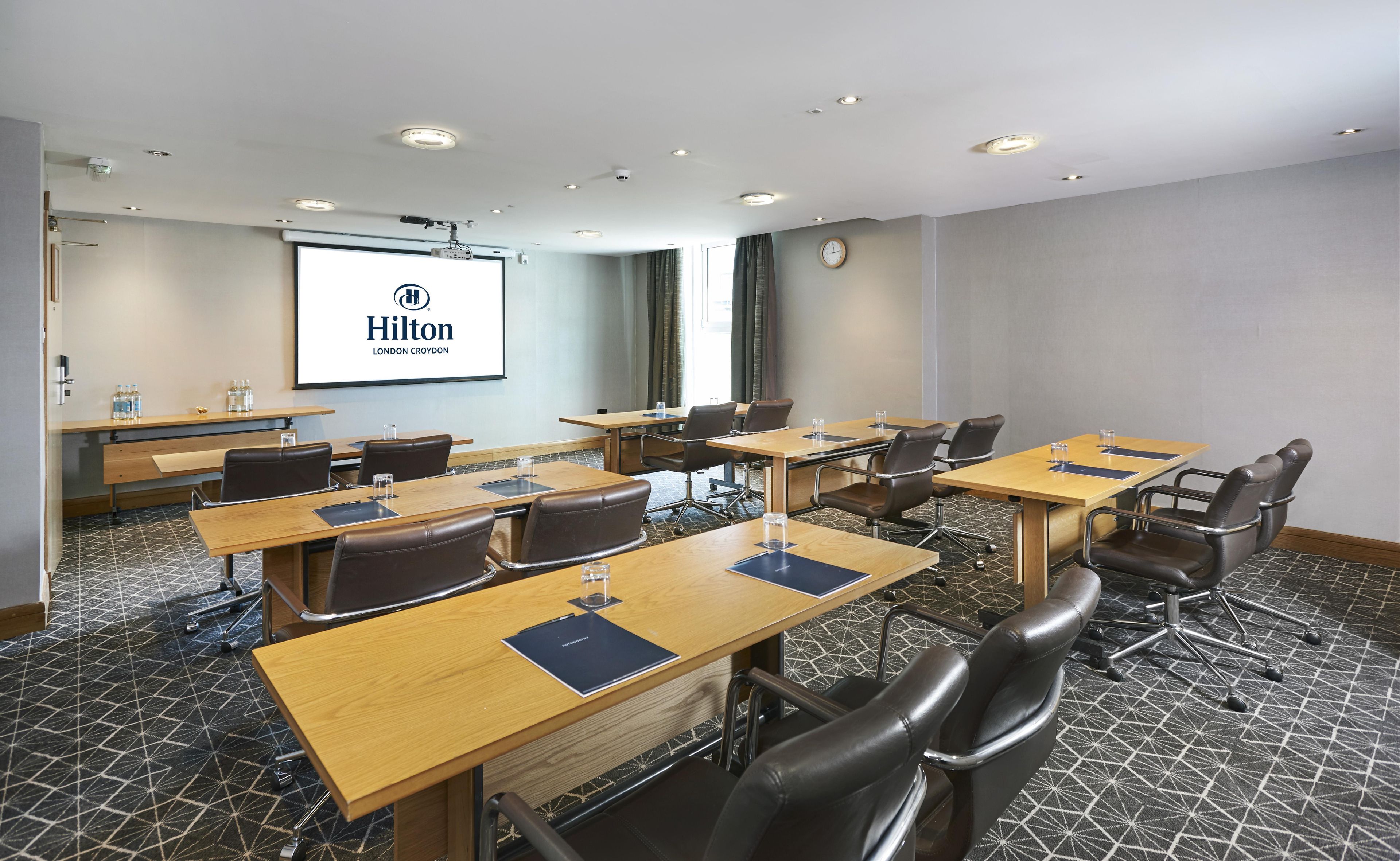 Une salle de conférence équipée de plusieurs tables, de chaises, d’un écran de projection affichant le logo de l’hôtel Hilton London Croydon, et d’une horloge au mur.