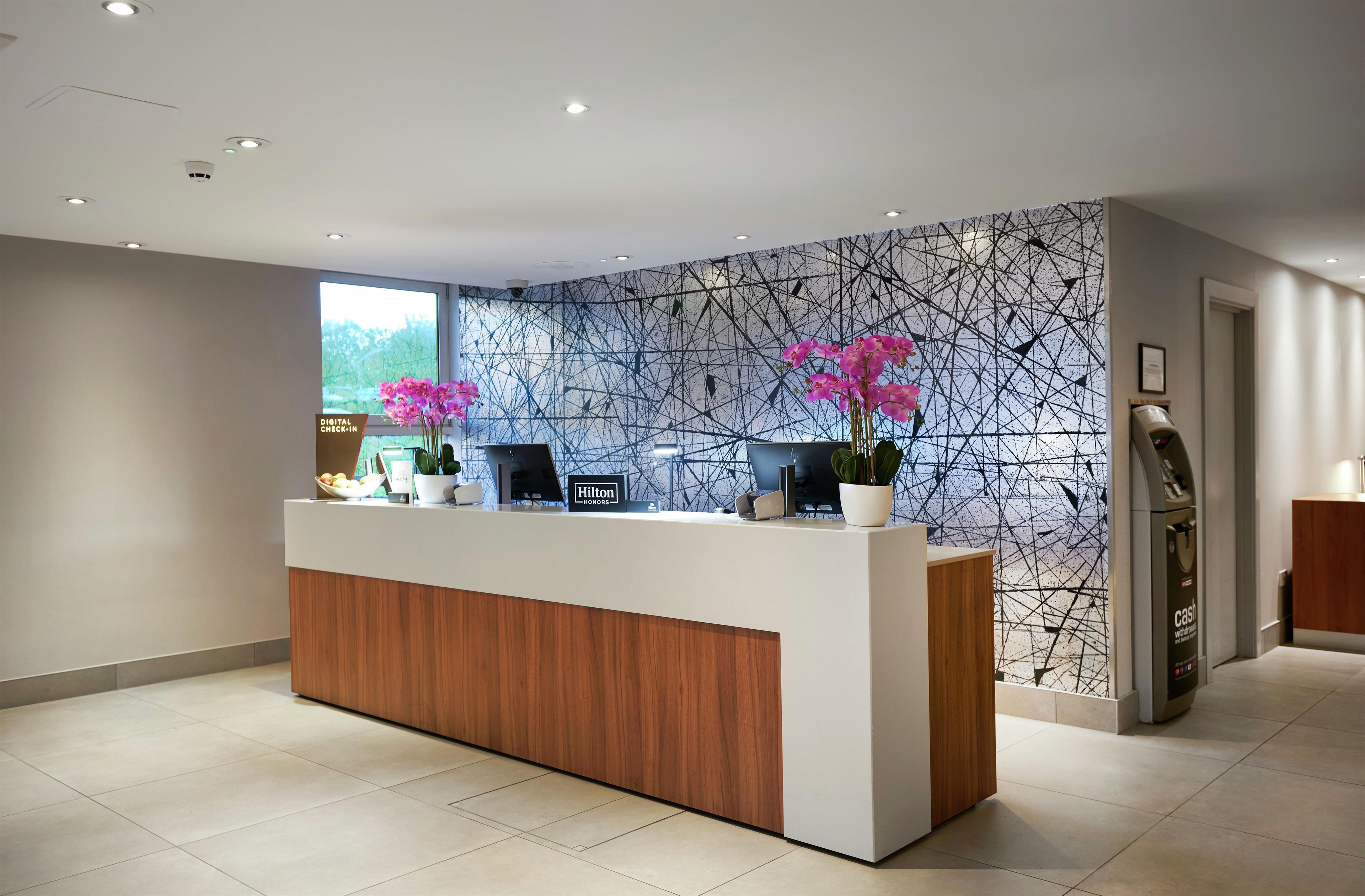 A hotel reception desk with a digital check-in station, a large window, and a wall with a geometric pattern.