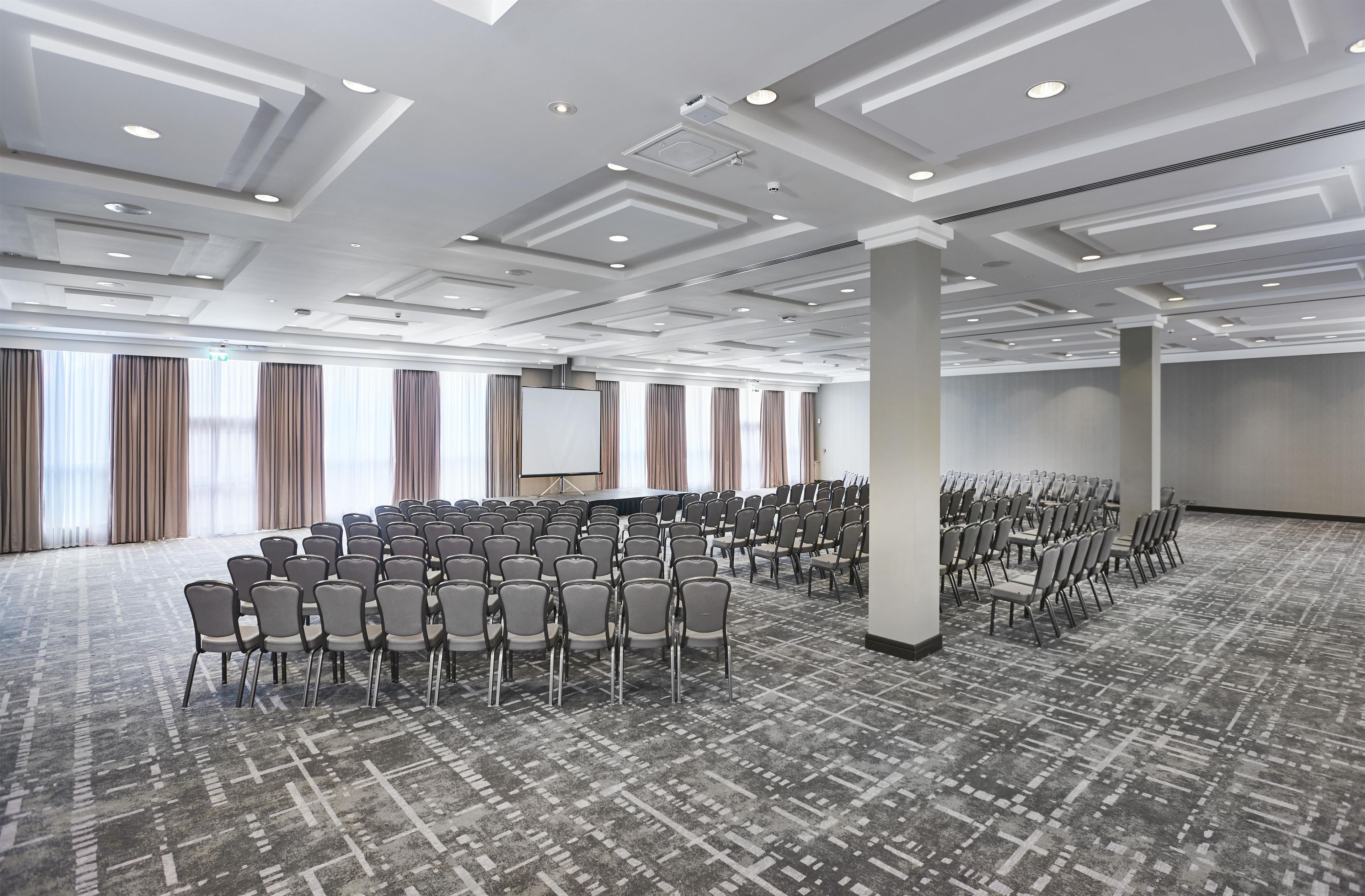 Une grande salle de conférence avec des rangées de chaises, un écran de projection et un sol recouvert de moquette.