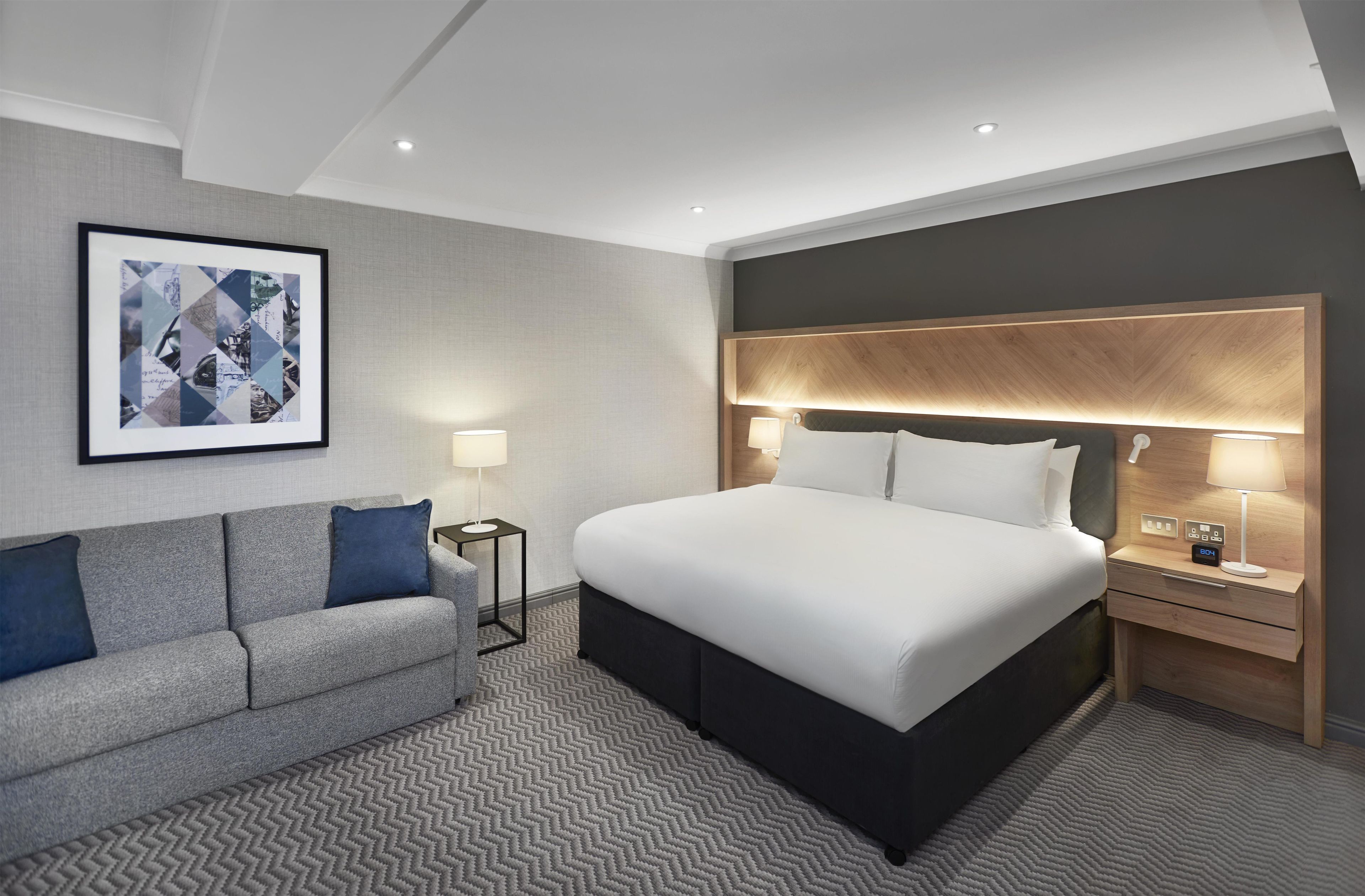 A hotel room with a bed, a sofa, two bedside tables, a wall-mounted artwork, and a patterned carpet.