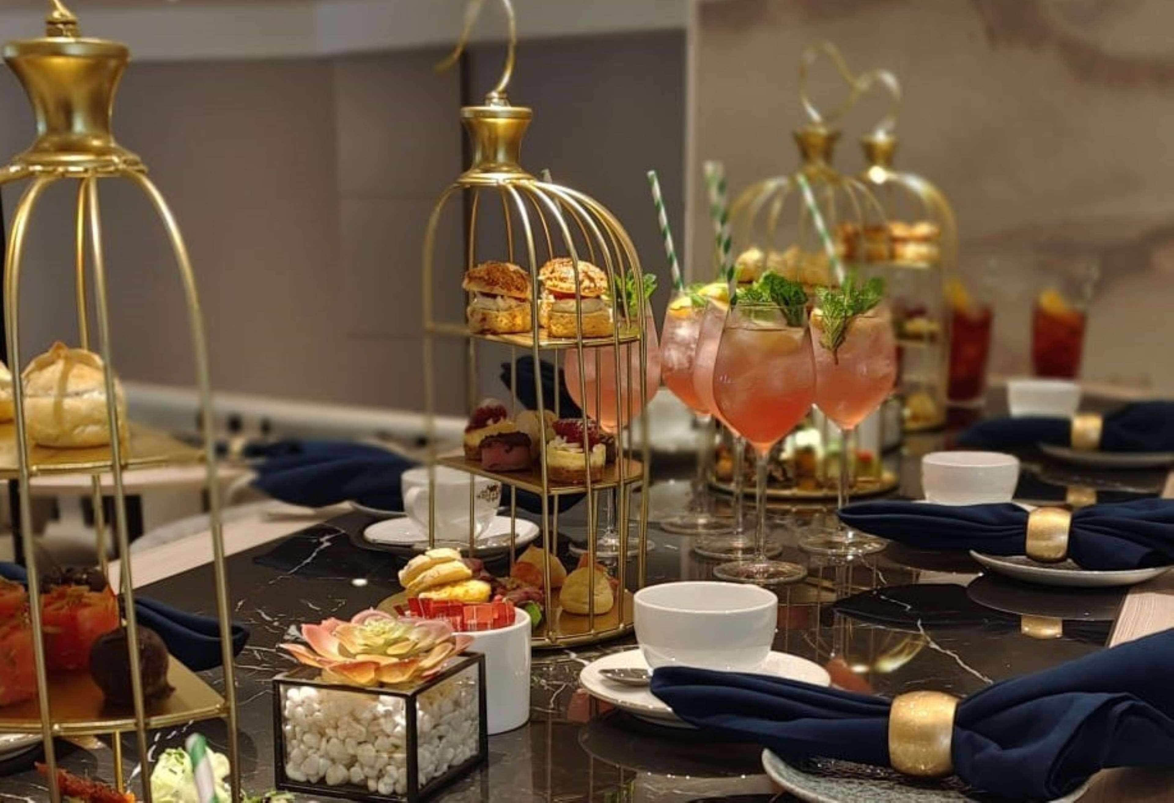 A table set with various desserts in glass containers, a pink cocktail, and a gold-colored birdcage.