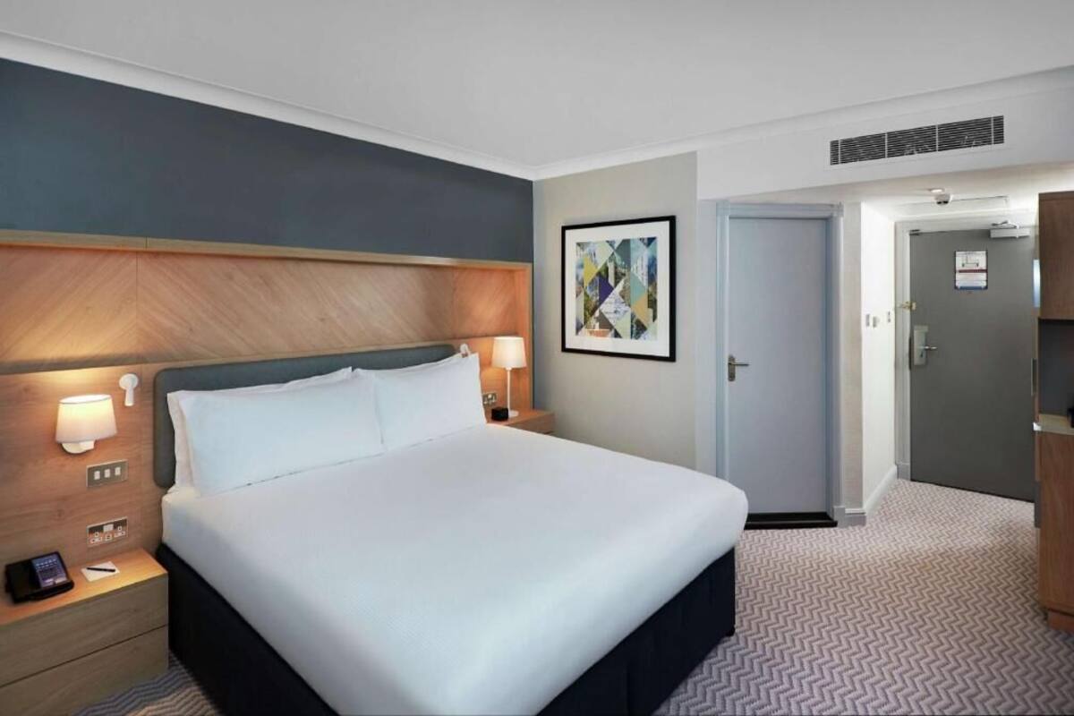 Habitación de hotel con cama, lámparas de noche, mesita de noche, una puerta y una obra de arte colgada en la pared.