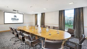 A conference room with a long table, chairs, a projector screen, and a view of greenery outside.