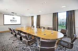 A conference room with a long table, chairs, a projector screen, and a view of greenery outside.