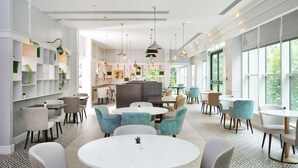 A modern interior with round tables and chairs, a kitchen area, and large windows providing natural light.