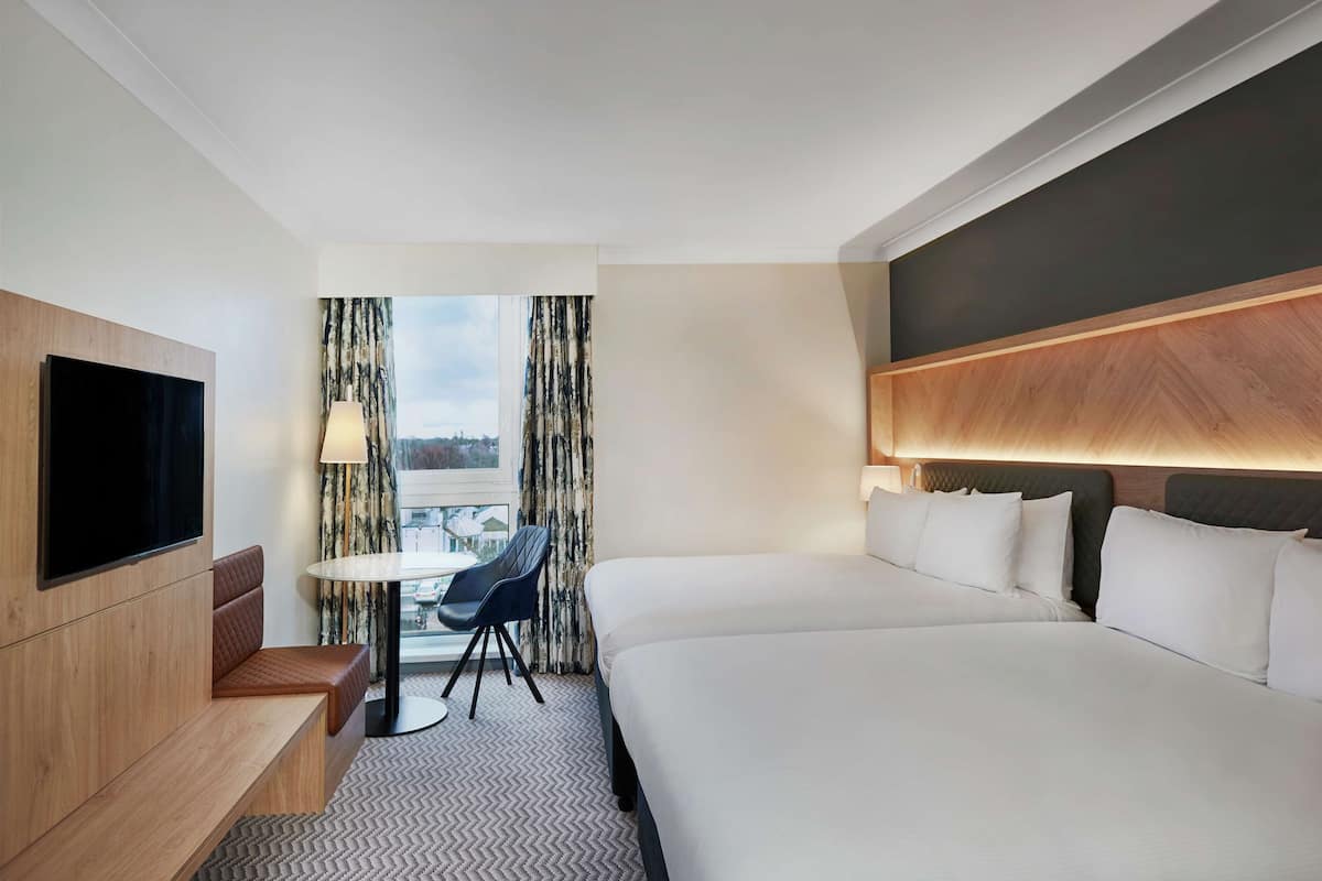 Habitación de hotel con una cama grande, un televisor montado en la pared, un área de estar con una mesa y sillas, y vista a la ciudad desde la ventana.