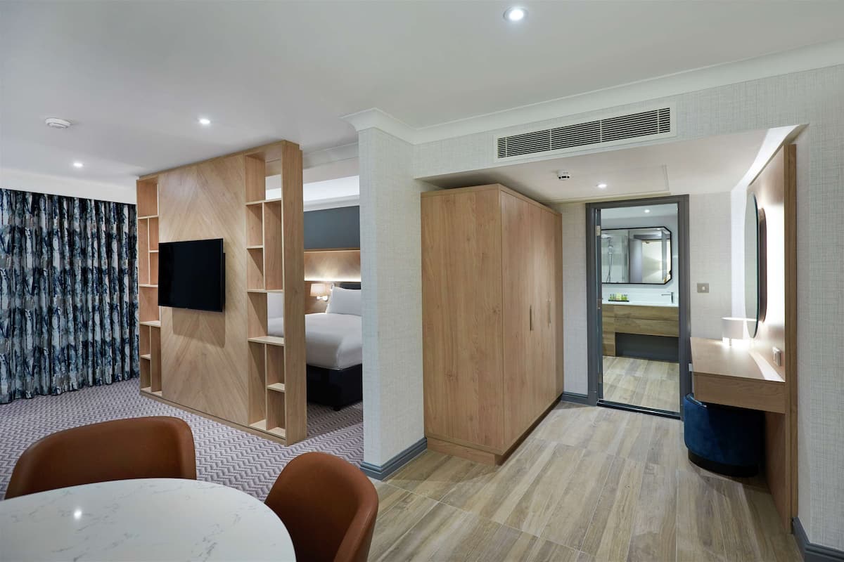 Habitación de hotel moderna con televisión de pantalla plana, armario de madera y baño con espejo y lavamanos.