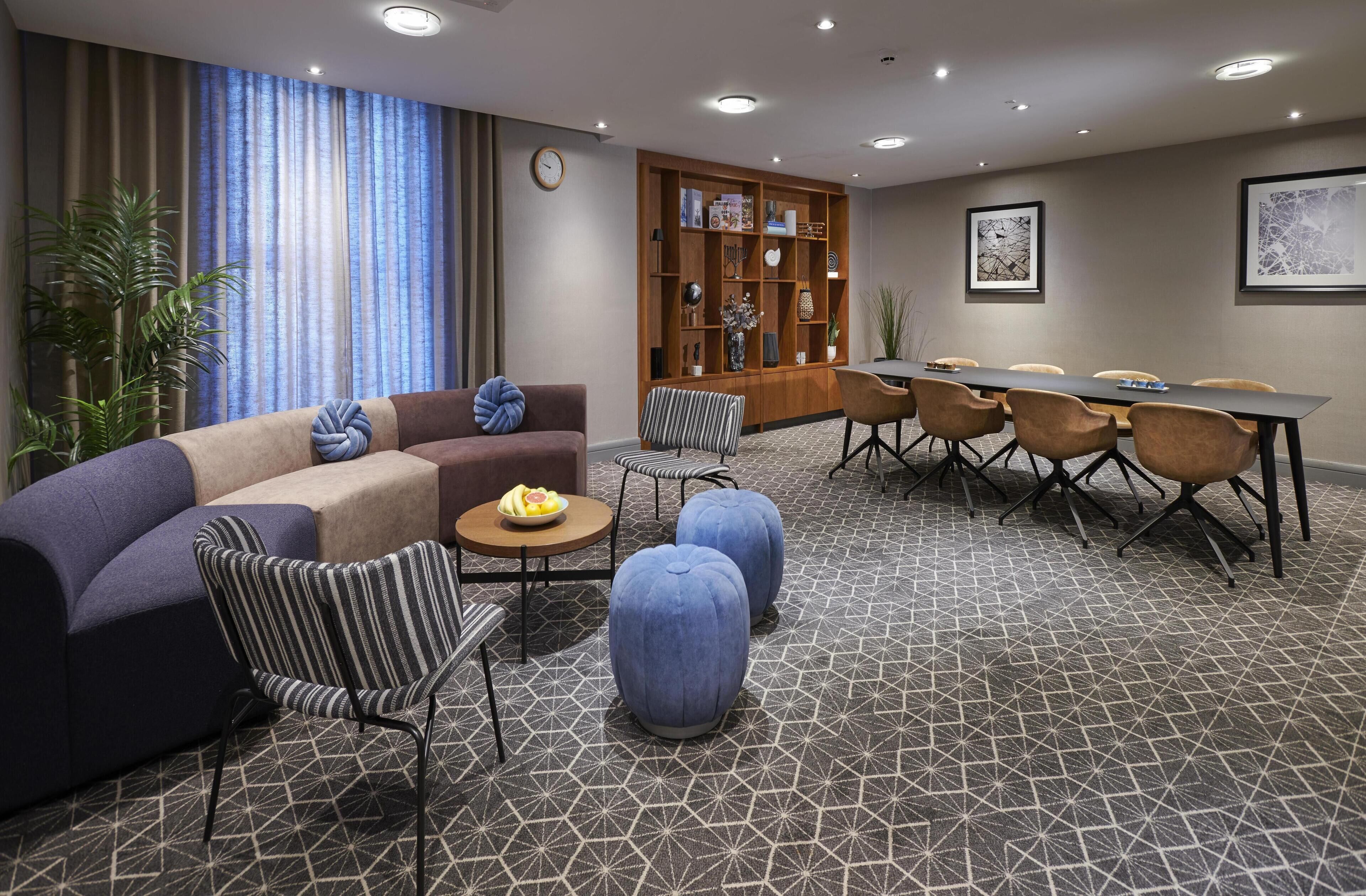 A modern hotel room with a sofa, armchairs, ottomans, a dining table with chairs, a bookshelf, and a large window with curtains.