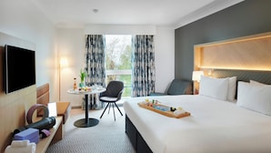 A hotel room with a large bed, a flat-screen TV, a desk with a chair, a small table with a lamp, and a window with curtains.