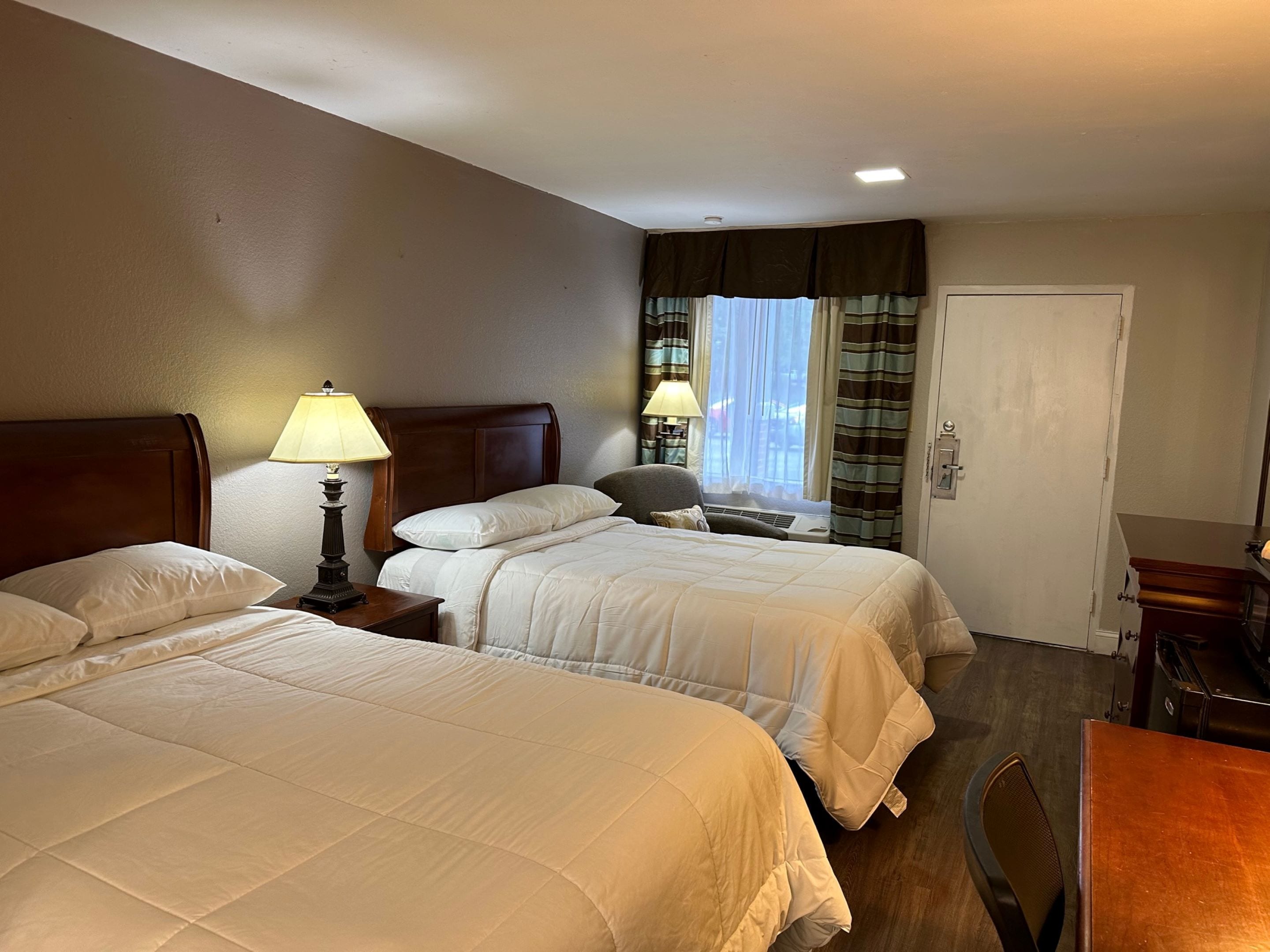 Une chambre d’hôtel avec deux lits, un bureau, une chaise, une lampe et une fenêtre avec des rideaux.