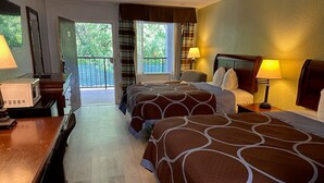 A hotel room with two beds, a desk with a microwave, a balcony with a view of greenery, and a door to the outside.