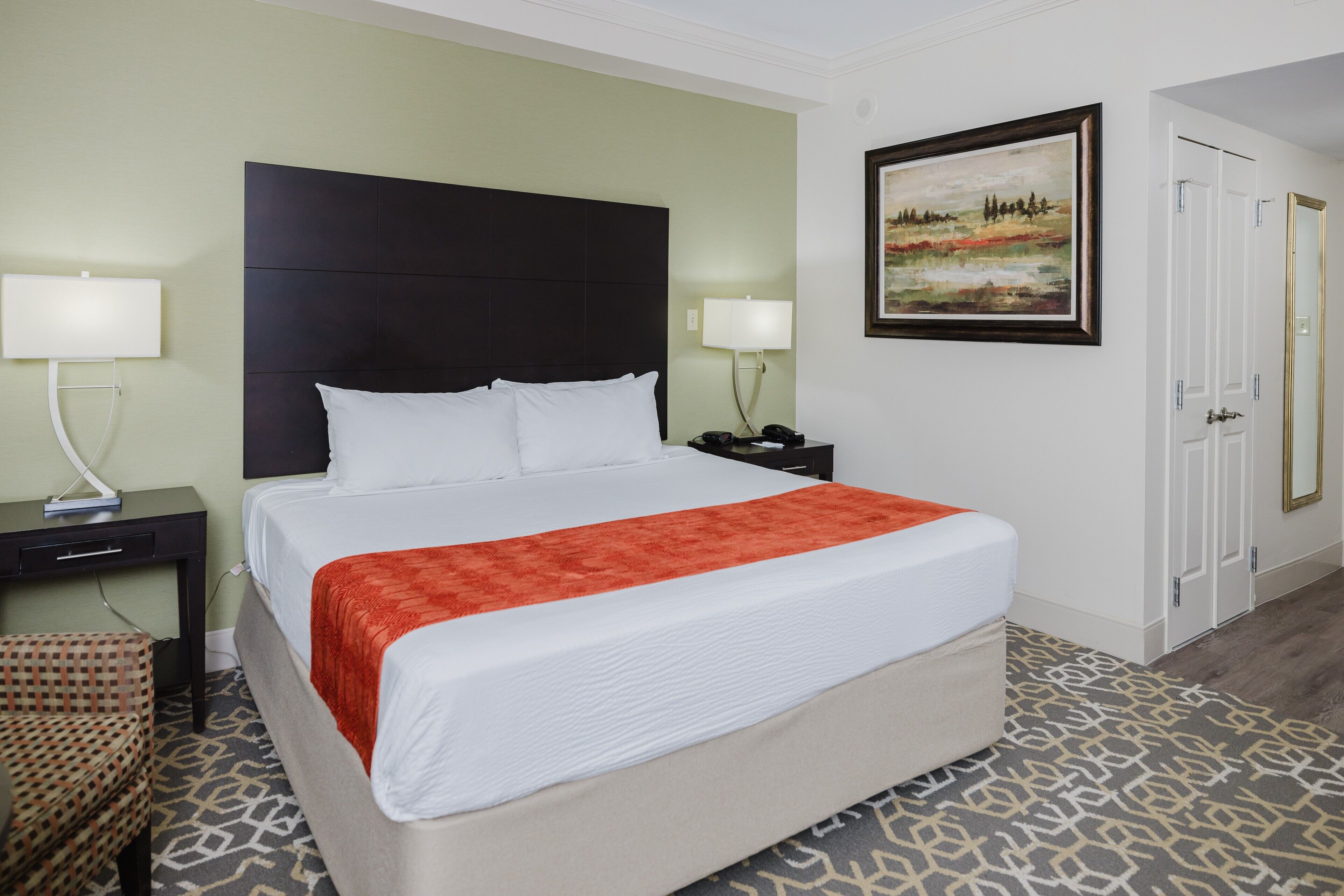 A hotel room with a large bed, two bedside tables, a chair, a painting on the wall, and a door.