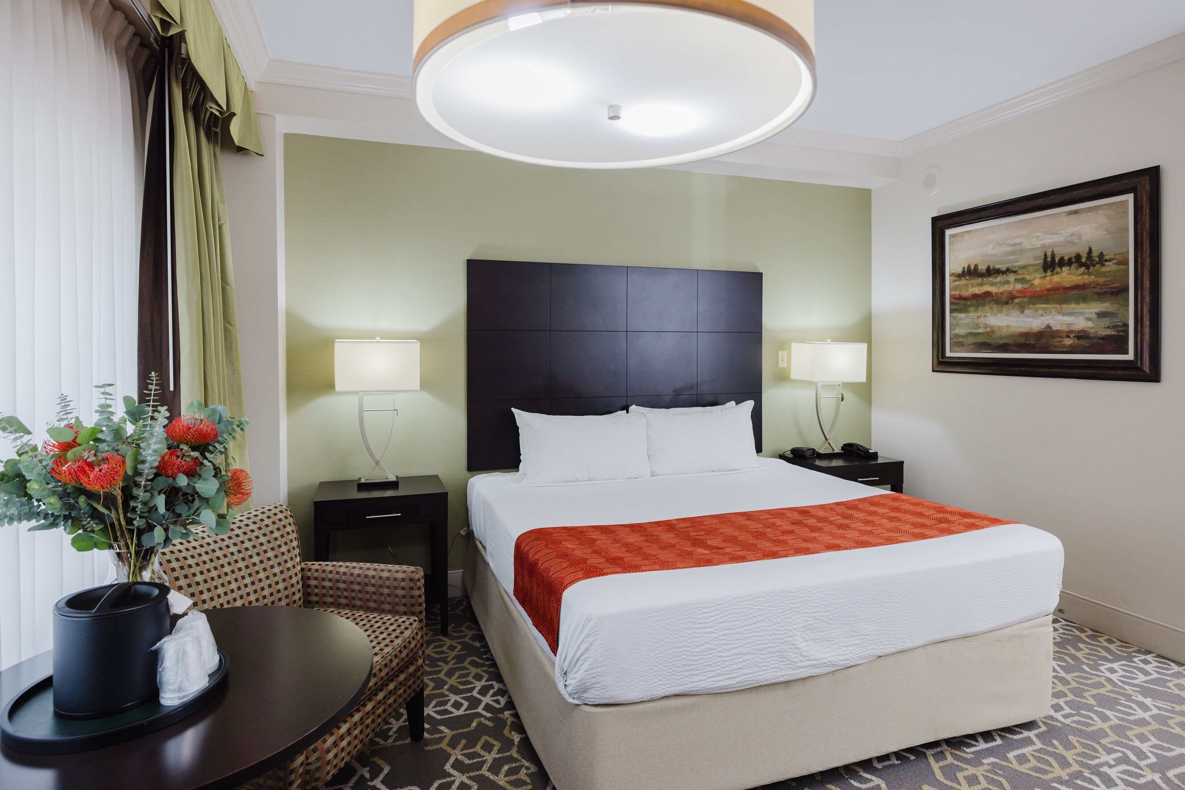 Habitación de hotel con una cama grande, dos mesitas de noche, una pequeña mesa redonda con un florero y un cuadro en la pared.