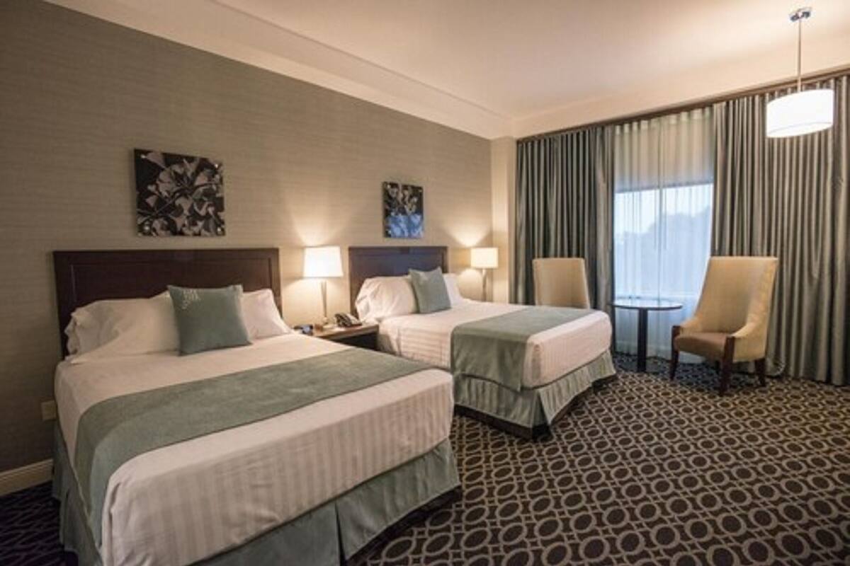 Hotellihuone, jossa on kaksi sänkyä, pieni pöytä, kaksi tuolia ja suuri ikkuna, jossa on verhot.