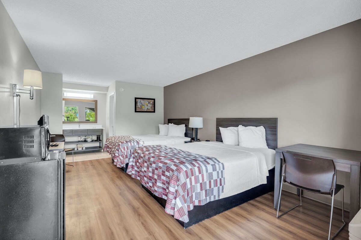 A hotel room with two beds, a desk, a chair, a refrigerator, and a window with a view of greenery.