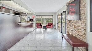 A hotel lobby with a reception desk, two red chairs, a bench, and a large window offering a view of the outdoors. - Red Roof Inn MacClenny (MacClenny)