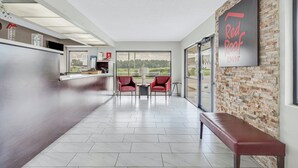 A hotel lobby with a reception desk, two red chairs, a bench, and a large window offering a view of the outdoors. - Red Roof Inn MacClenny (MacClenny)
