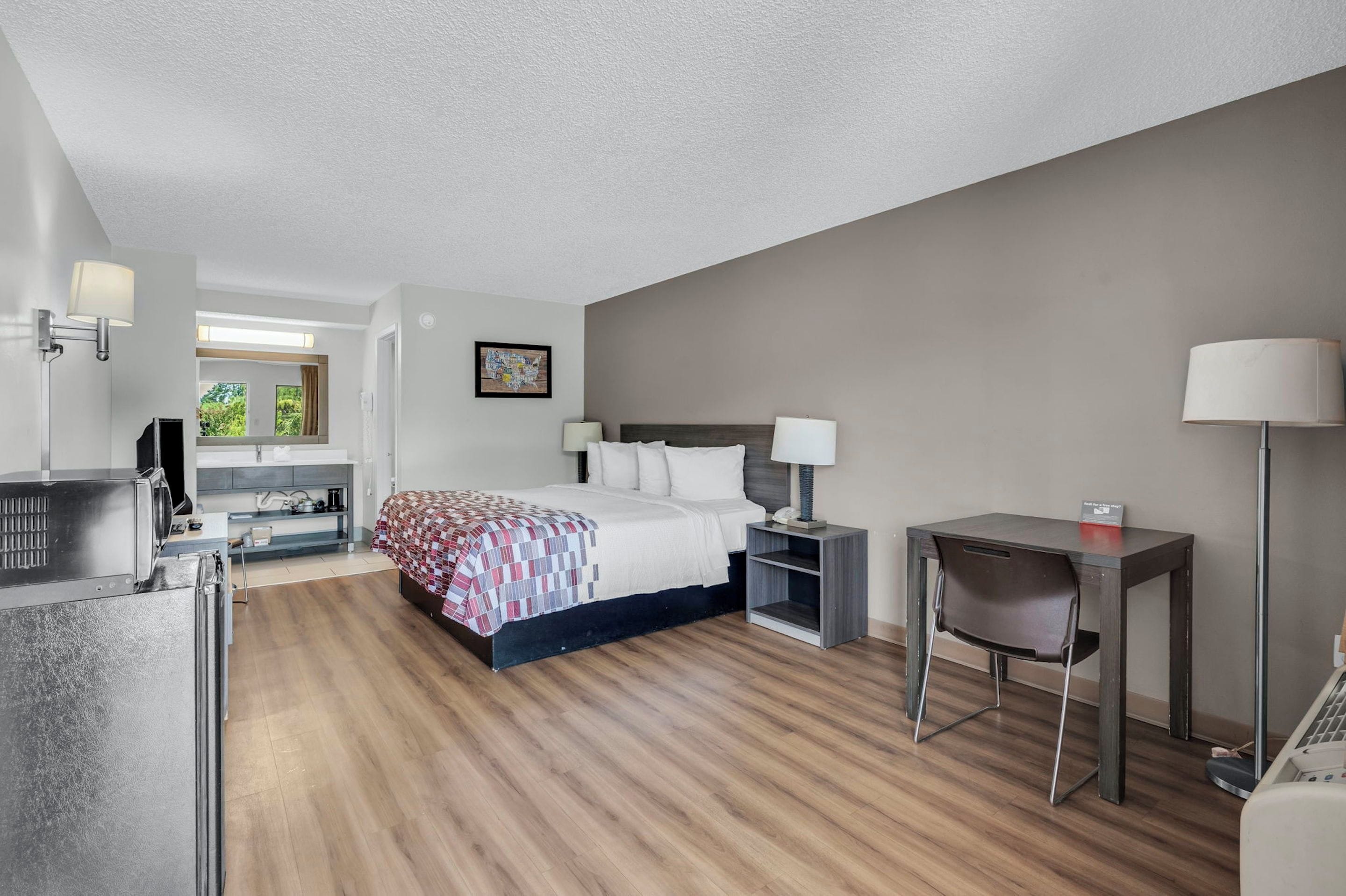 A hotel room with a bed, bedside tables, a desk, a chair, and a view of greenery outside.
