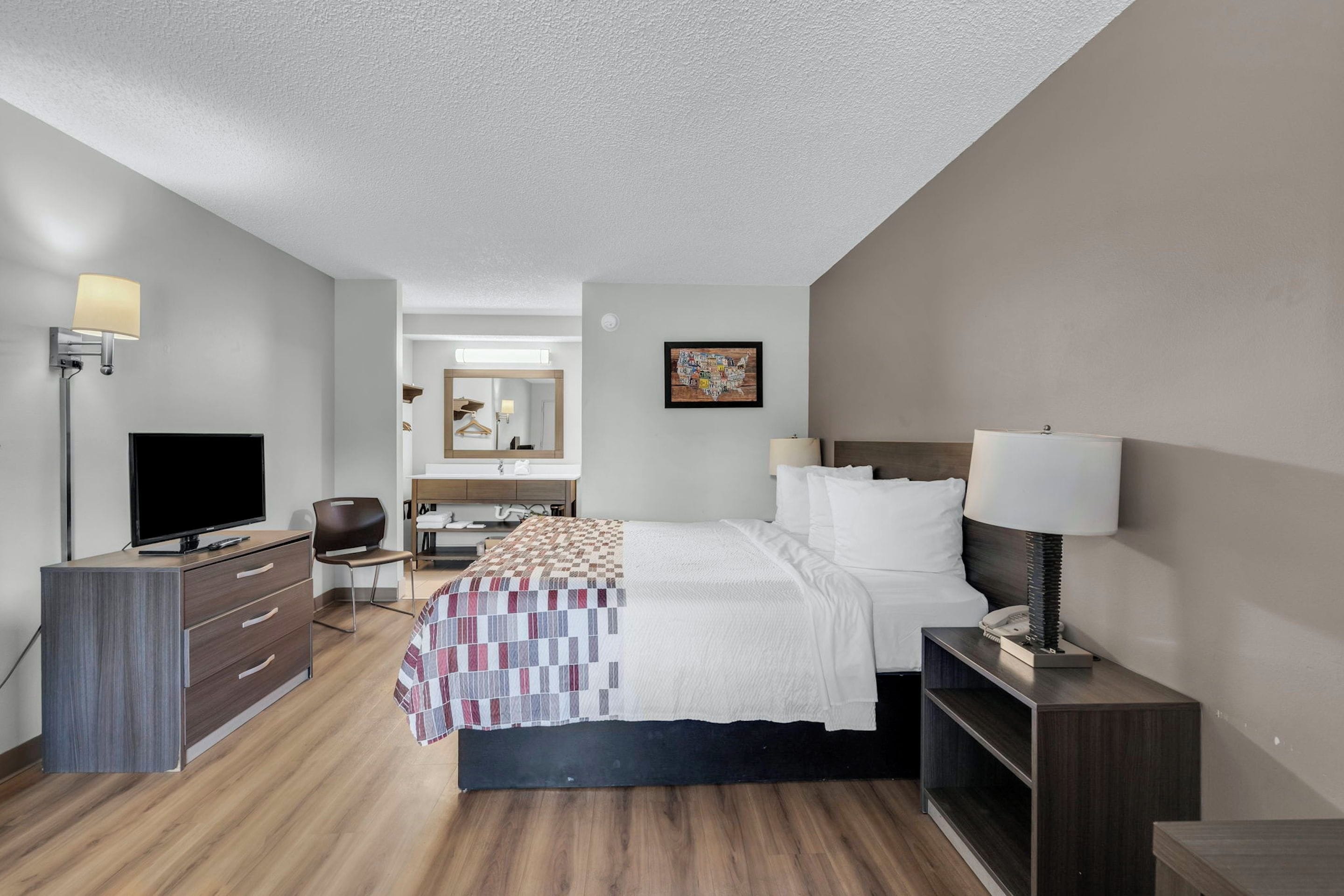 Habitación de hotel con cama, mesita de noche, lámpara, televisor, cómoda y espejo.